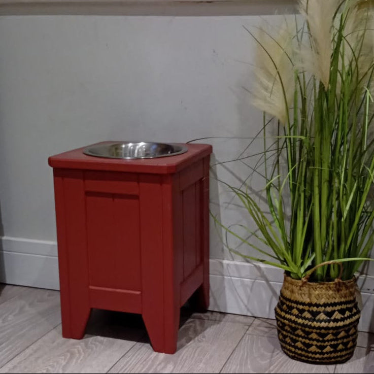 37cm tall handmade wooden raised dog bowl painted in farrow and ball eating room red with a stainless steel bowl and a tall grass plant next to it on a laminate floor against a neutral background