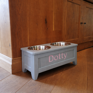 grey wooden raised dog bowl with pink personalised name on the front that reads Dotty with a wooden floor and wooden kitchen cupboards in the background