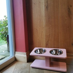 Pink pet feeder with two bowls on a wooden floor next to a window.