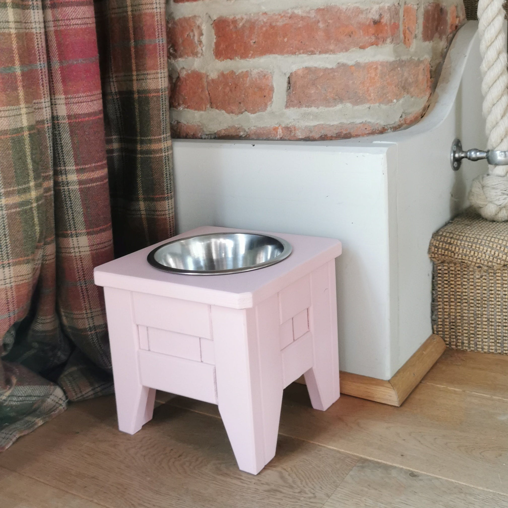 Pink handmade raised wooden stand with a metal bowl on a wooden floor next to plaid curtains and a brick wall.