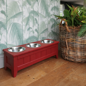 handmade wooden triple raised dog bowl painted in farrow and ball eating room red, 27cm high with three stainless steel bowls on a wooden floor with a leafy wallpaper behind and a green plant in a wicker basket
