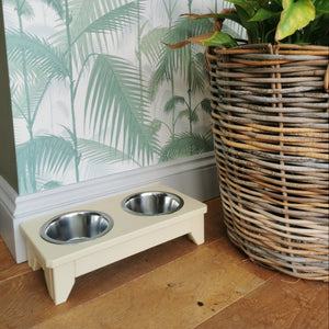 Handmade raised wooden double dog bowl painted in farrows cream  with two stainless steel bowls on a wooden floor next to a large wicker basket with plants.