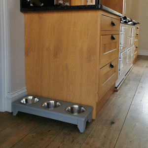 Kitchen with wooden cabinets, a white aga, and a handmade raised wooden triple dog bowl stand painted in grey on a wooden floor. 
