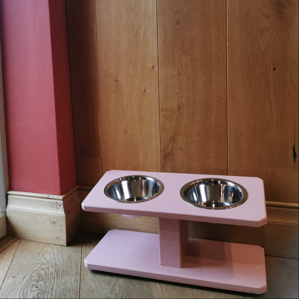 Pink pet feeder with two bowls on a wooden floor next to a window.